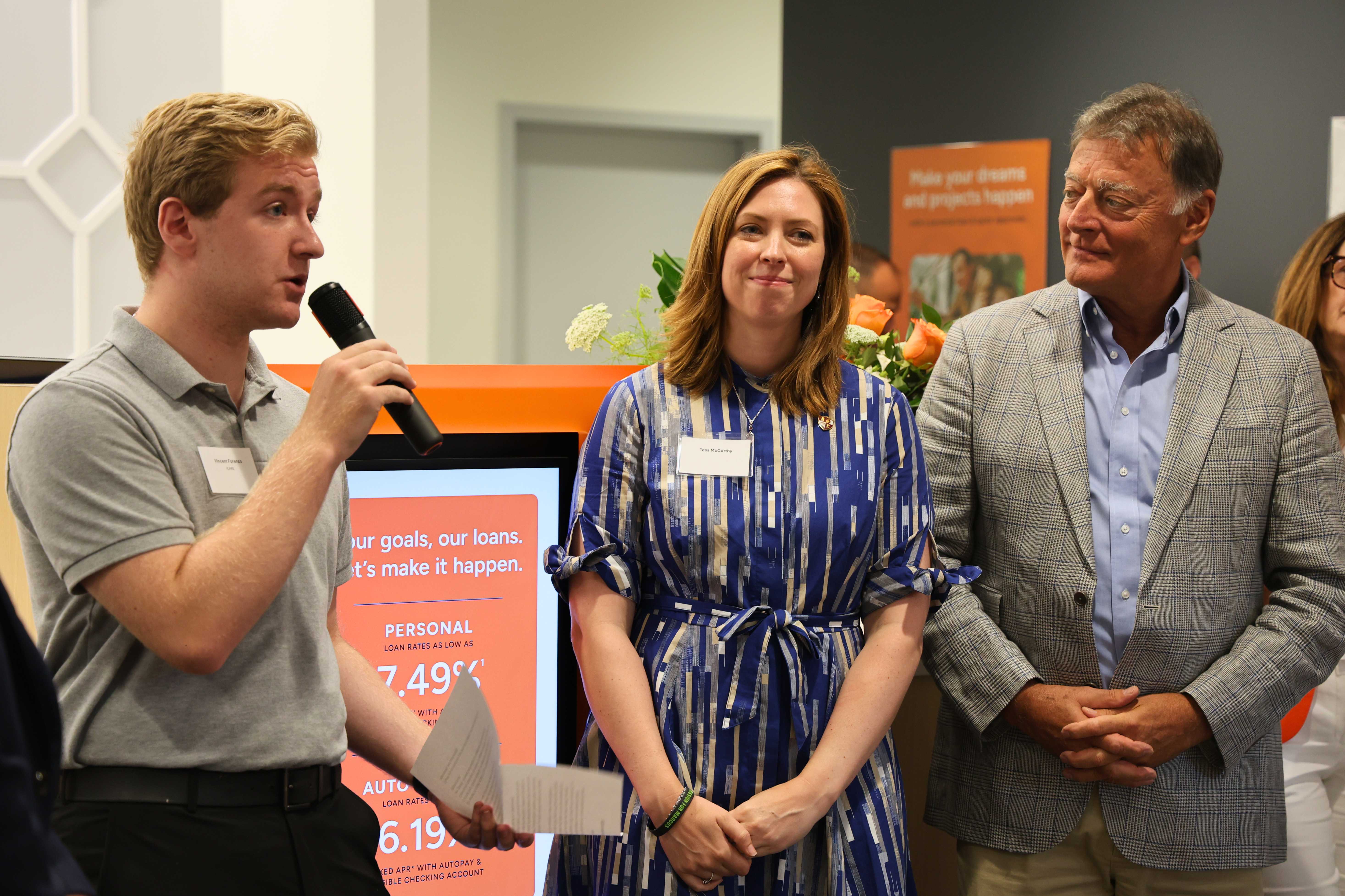 A student stands in front of a room holding a microphone and speaking to the crowd while a man and woman look on smiling